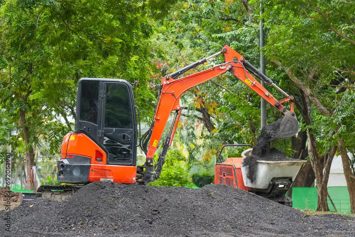 Orange and black compact excavator with a full bucket of dark aggregate.