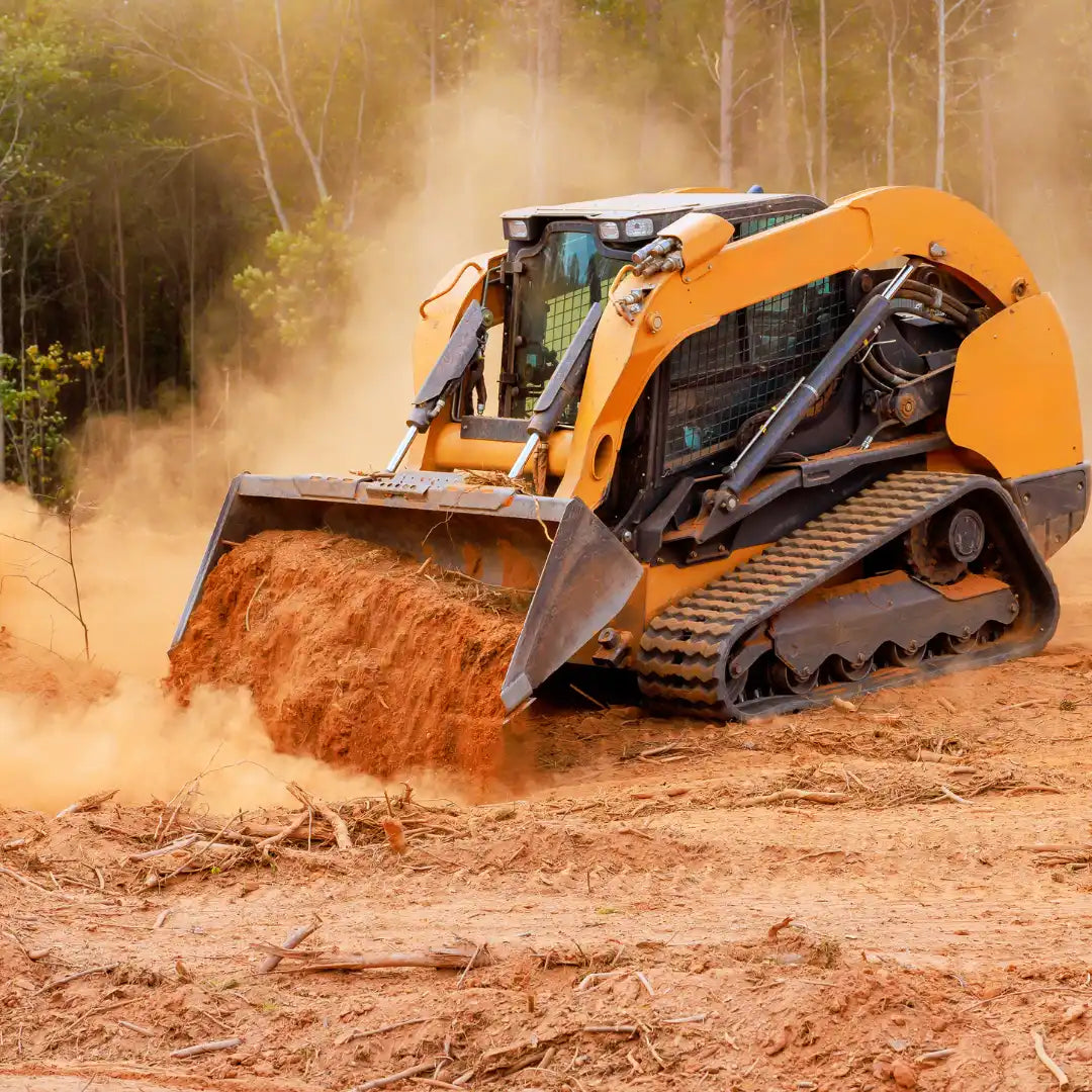 Yellow skid steer loader operating in a forest clearing with dust and trees in the background.