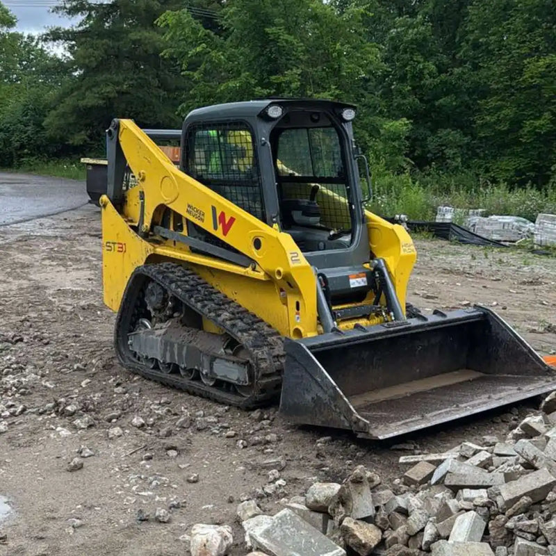 A Wacker Neuson ST31 compact track loader, featuring 13-inch wide rubber tracks and a front bucket, positioned next to a pile of concrete debris on a jobsite under an overcast sky
